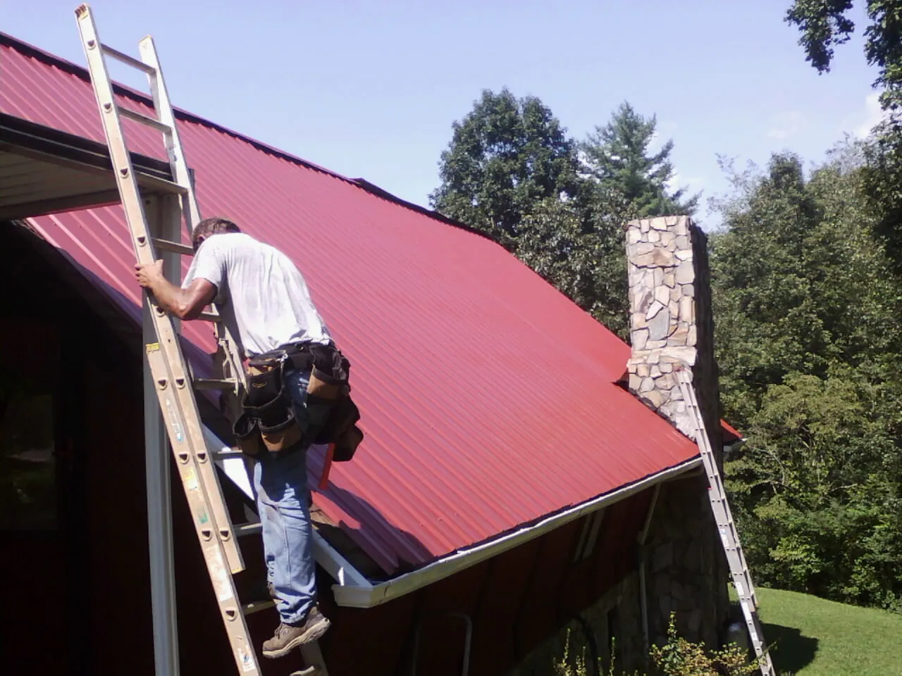 Roofer on a ladder beside a red metal roof and stone chimney — Soffit & Fascia Repair by Roofing Brothers in Lanham